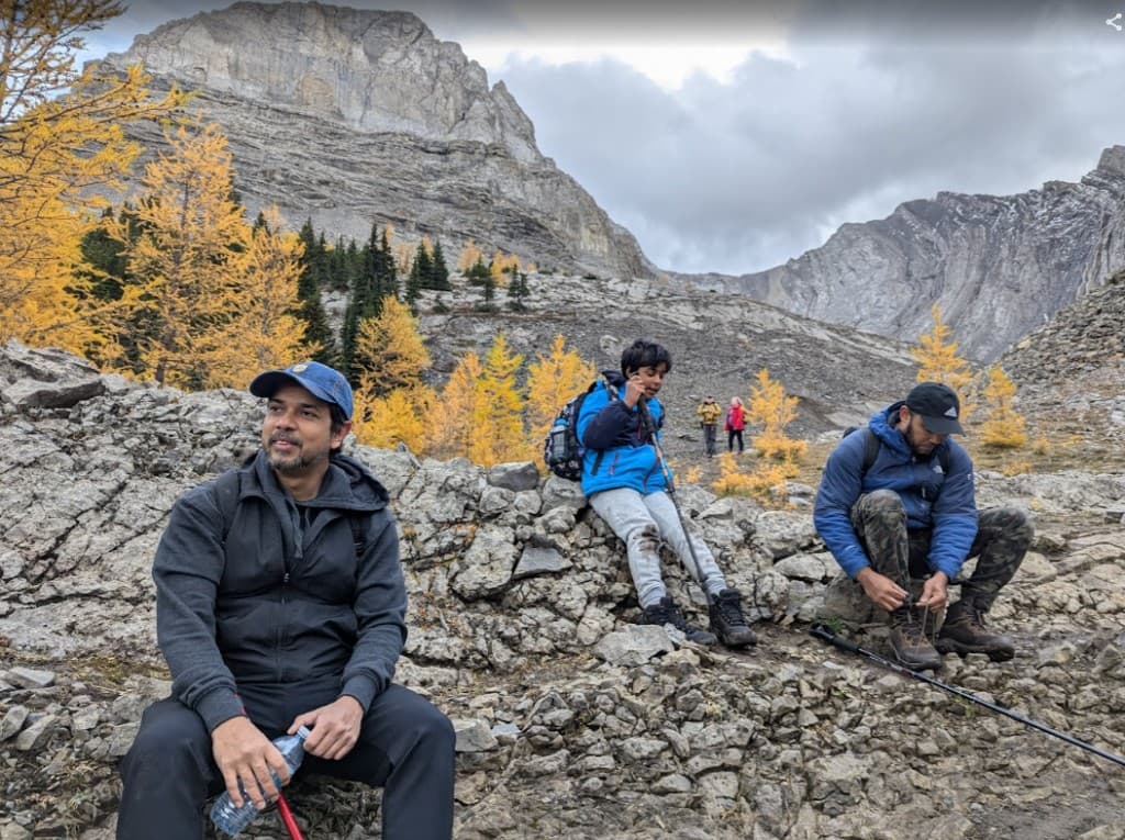 Hikers taking a break on a rocky trail with golden larch trees and mountain cliffs at Arethusa Cirque