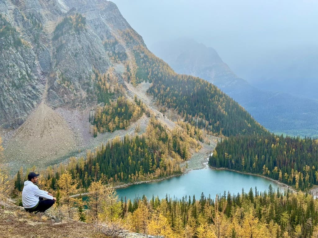 Hiker on a rocky ledge overlooking a turquoise alpine lake surrounded by golden larch forest and misty mountain peaks