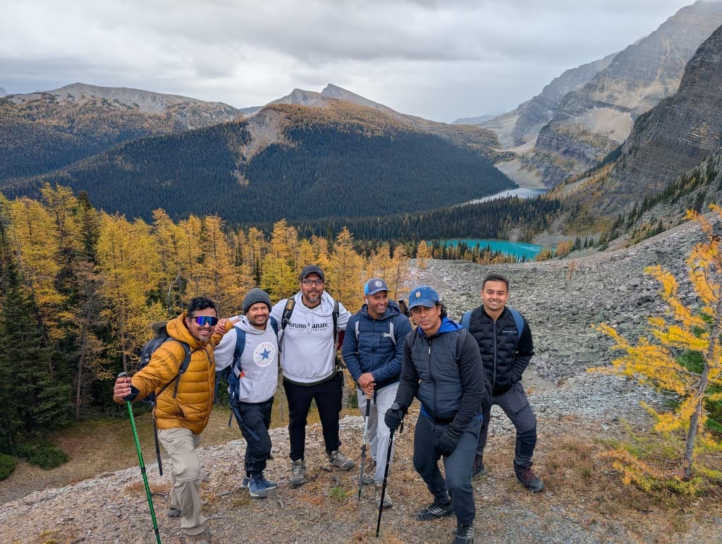 Six hikers posing on a rocky ridge with turquoise alpine lakes and golden larch trees in the valley behind them