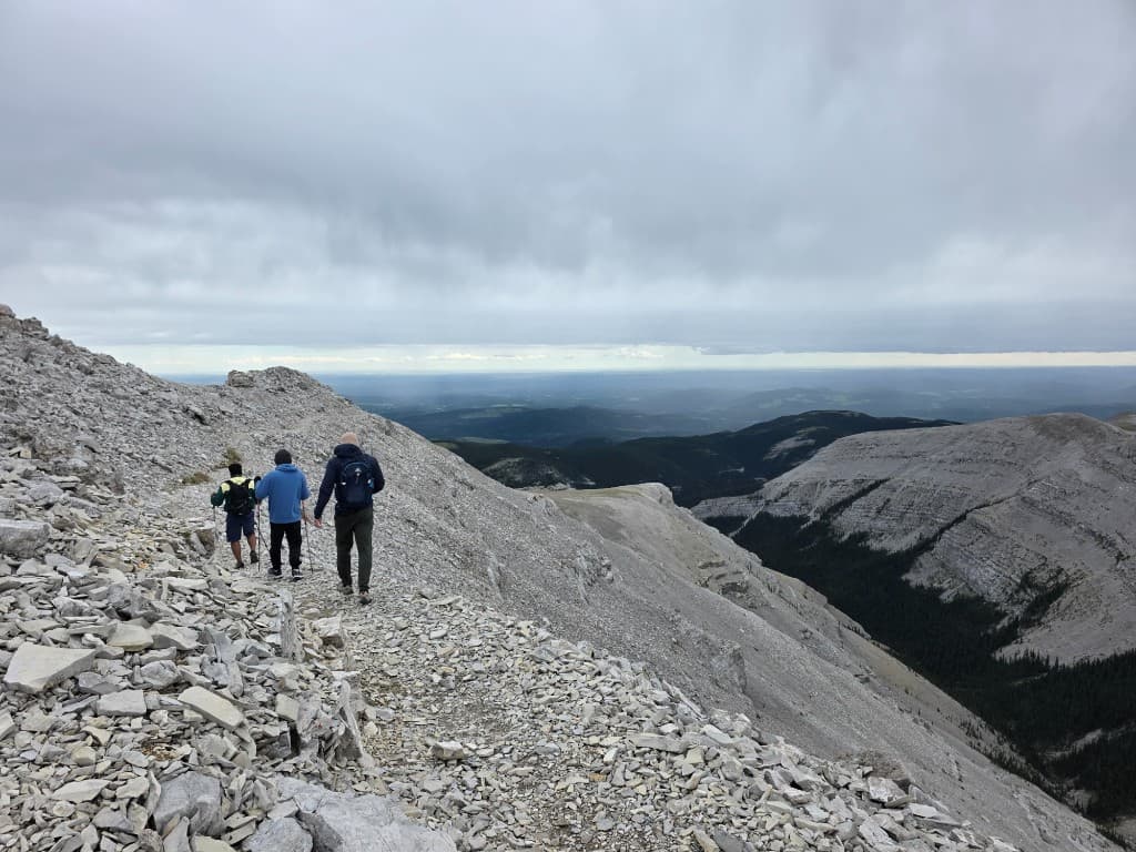 Three hikers with backpacks and trekking poles on a steep rocky ridge overlooking a forested valley under a cloudy sky