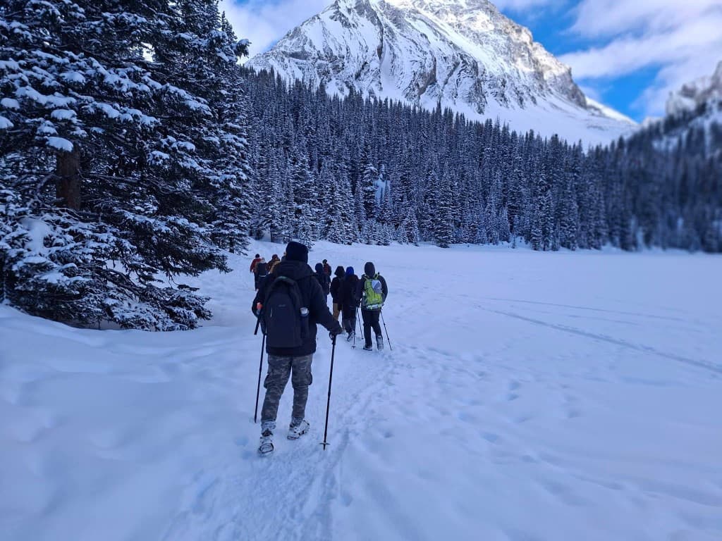 Line of hikers trekking across a snow-covered frozen lake toward evergreen forest and a snow-capped mountain peak