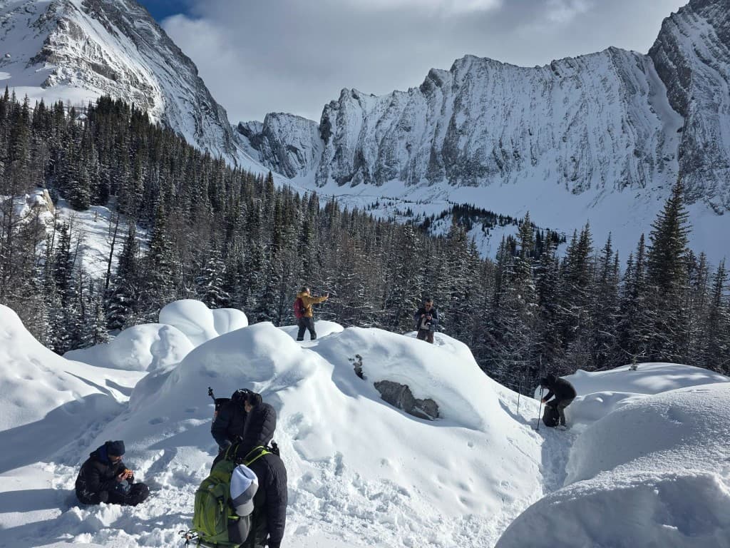Hikers on deep snow mounds with a snow-covered mountain ridge and evergreen forest in the background
