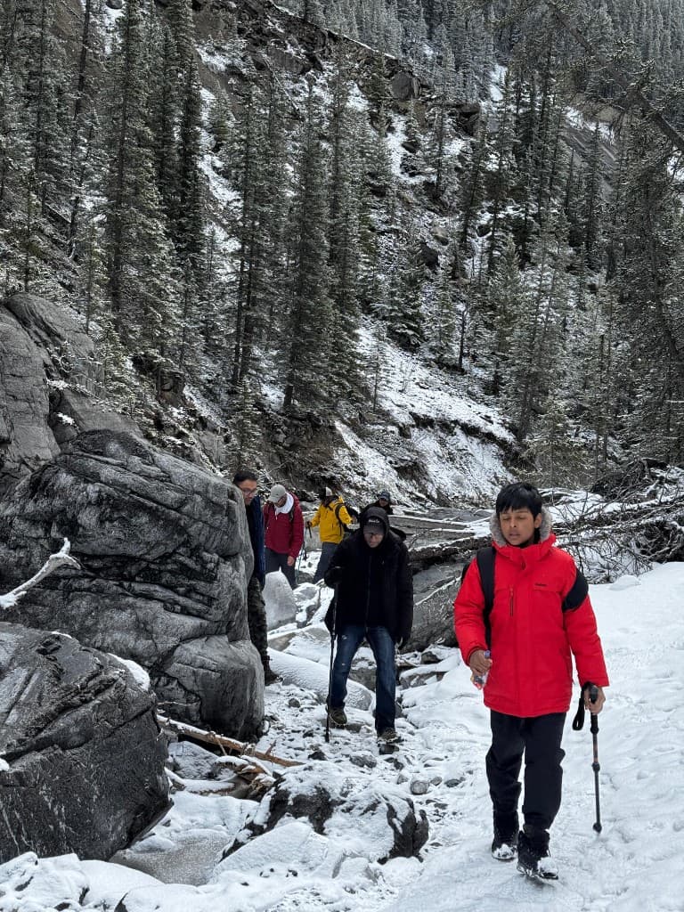 Hikers on a snowy rocky trail through a dense evergreen forest, led by a person in a red winter parka