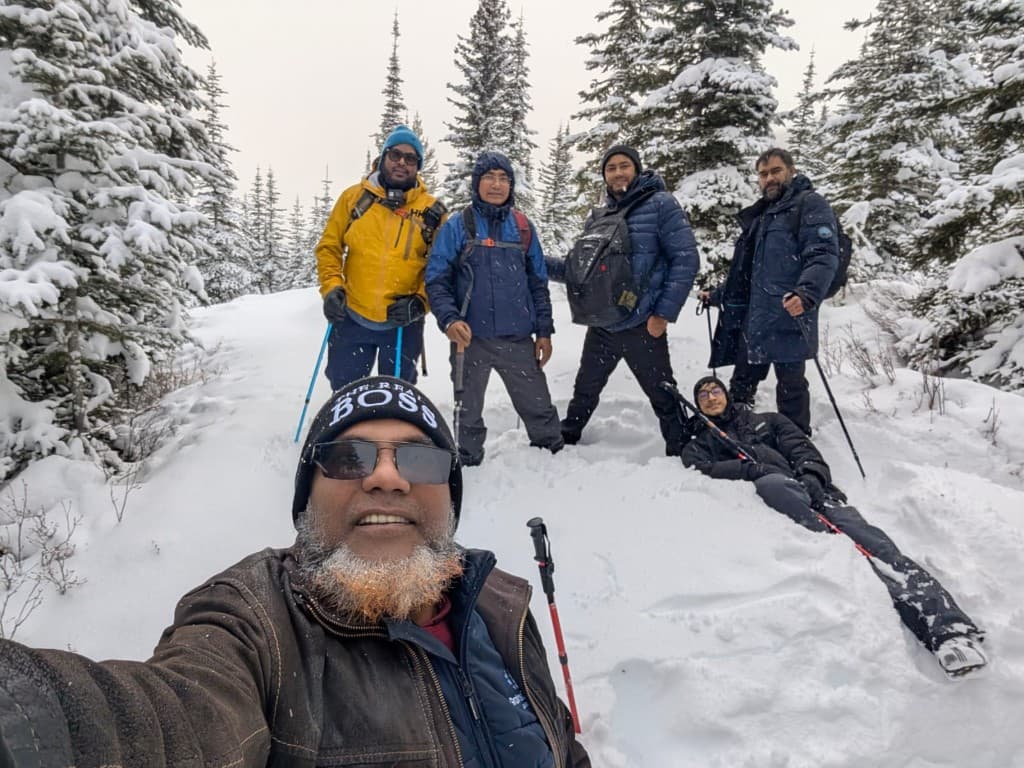 Six hikers posing for a group selfie in a snow-covered evergreen forest, with winter jackets, backpacks, and trekking poles