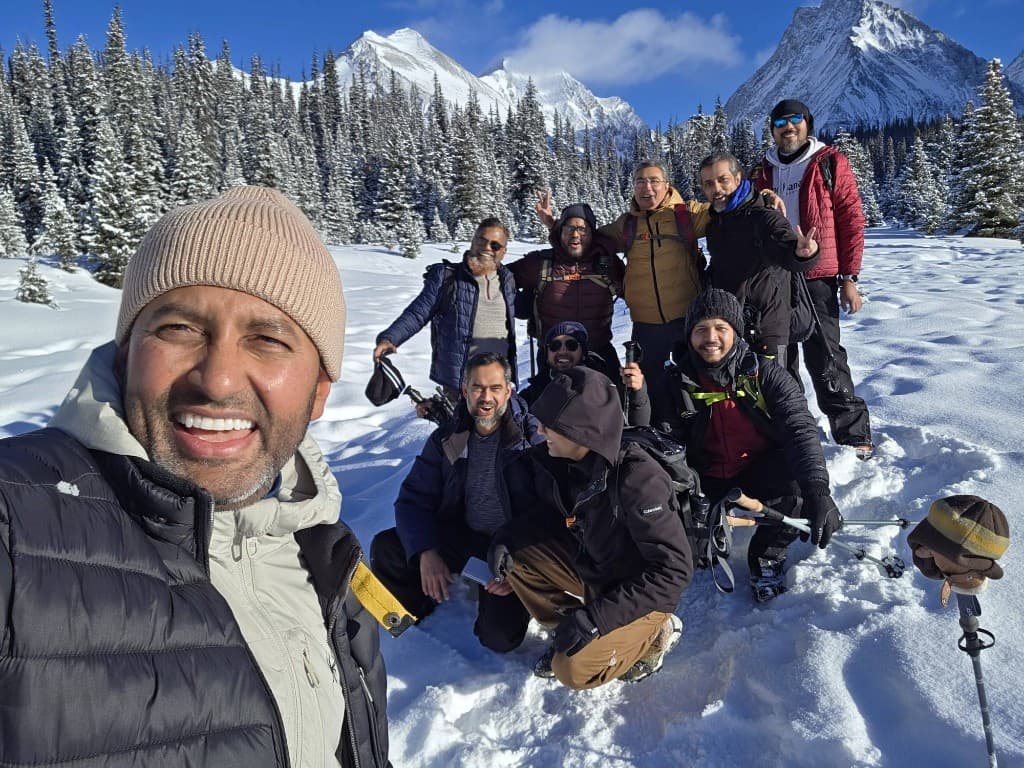 Group of hikers smiling in deep snow with snow-covered evergreens and mountain peaks under a bright blue winter sky